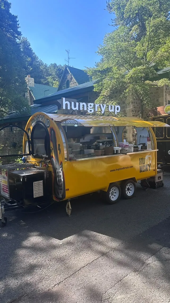 The yellow Hungry Up trailer open and ready to serve on a pine-shaded road in a mountain village, rooftop sign above the hatch.